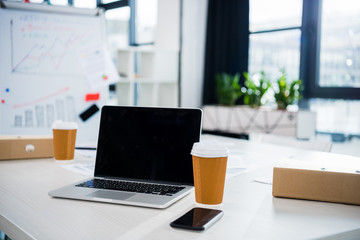 laptop computer, smartphone and coffee cups on workplace in empty office, small business office