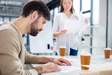 Businesswoman bringing papers to bearded young businessman looking at contract at workplace