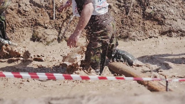 A group of active young people in camouflage pants running and crawling through mud pit in obstacle endurance race. Slow motion