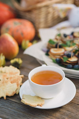 White cup and saucer with tea and cookies in the form of a maple leaf on a wooden table. Against a background of pumpkins and pancakes.