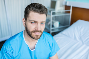 Portrait of sick bearded man sitting on hospital bed and looking at camera