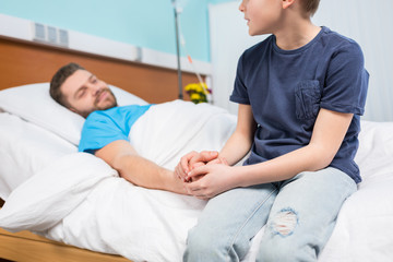 Fototapeta premium Cropped shot of son holding hand of sick father lying in hospital bed, dad and son in hospital