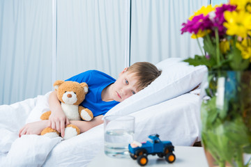 Sick little boy hugging teddy bear and lying in hospital bed