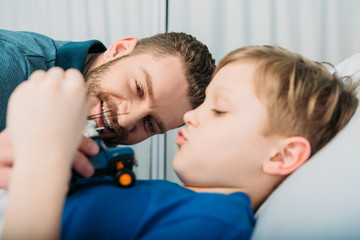 Smiling father playing with sick little boy lying in hospital bed, dad and son in hospital