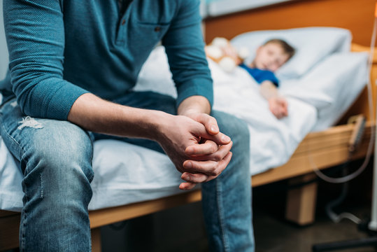 partial view of dad sitting near sick son in hospital bed