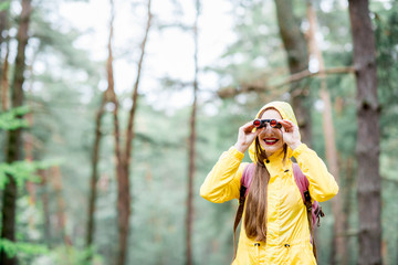 Young woman in yellow raincoat looking with binoculars in the green pine forest. Bird watching in...