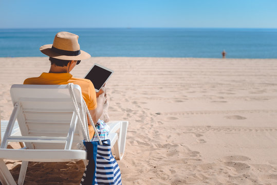 Back view of man sitting using touch screen tablet pc. Beach sunny seaside outdoors background. Travel lifestyle modern business connectivity, selfie photography, online shopping or playing game