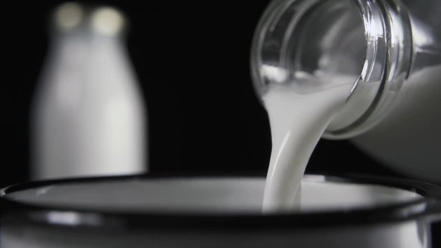 Slow motion. Milk from a glass bottle is poured into a mug in the foreground, behind is a full bottle of milk. Black background
