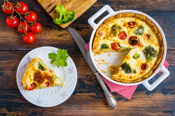 Homemade quiche with broccoli and cherry tomatoes in the baking dish and a piece of cake on a plate on the rustic wooden table, top view.