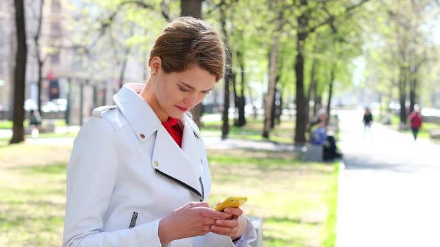 SMS. Young beautiful girl reads messages on cellphone sitting on bench in park