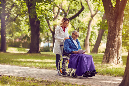 Elderly man reading book in wheelchair with nurse in the park.