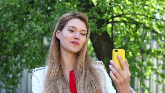 Video conference. Young beautiful woman communicates on cellphone sitting on bench in park