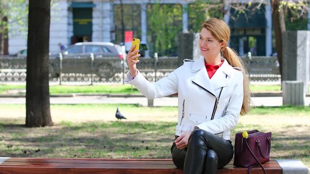 Video conference. Young beautiful girl communicates on cellphone sitting on bench in park