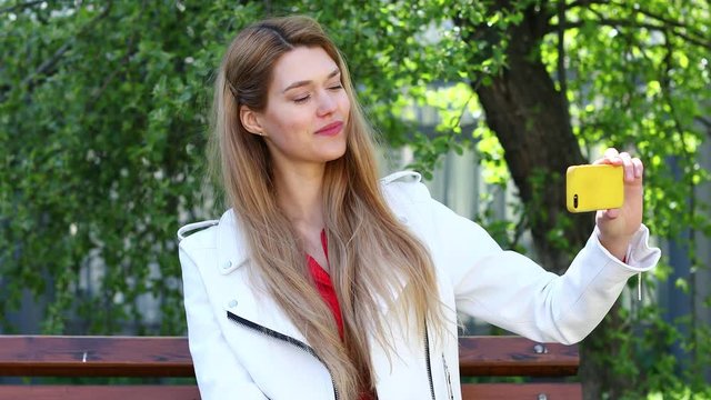 Video conference.  Beautiful blonde woman  communicates on cellphone sitting on bench in park