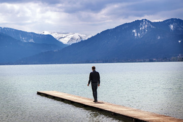 Fototapeta na wymiar Young man alone on a jetty in the tegernsee lake looks to the mountains, itourist resort of the Bavarian Alps
