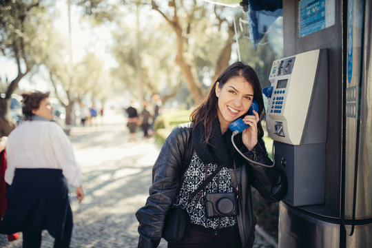 Smiling young woman speaking on a public payphone outside.Happy expression.Dialing local number in foreign country,low cost calls for international dials.Coin or card public phone used by tourist