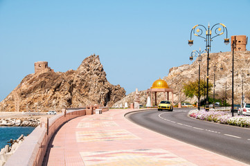 Ulica i deptak wzdłuż wybrzeża w Muscat z widokiem na ruiny fortów na skale.  - obrazy, fototapety, plakaty