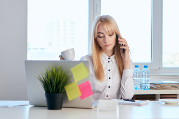business woman with a cup of coffee sitting at the computer, talking on the phone, a plant