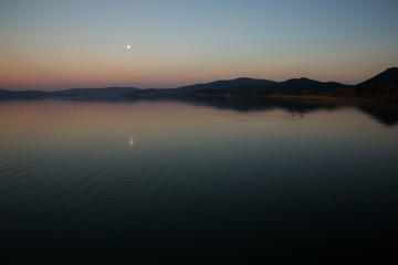 Fototapeta na wymiar A minimalist view of a dusk at a lake, with perfectly symmetric reflections, and small moon reflecting on water