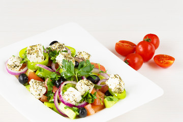 Greek salad with fresh tomato, cucumber, red onion, basil, feta cheese, black olives, Italian herbs and olive oil in white dish on white wooden table.