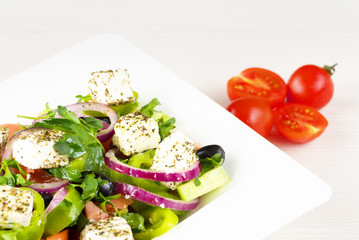 Greek salad with fresh tomato, cucumber, red onion, basil, feta cheese, black olives, Italian herbs and olive oil in white dish on white wooden table.