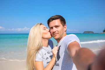 Fototapeta na wymiar Outdoor closeup portrait of pretty young couple in love takes a self-ie having fun in hot weather and feeling happy together on the tropical island. Posing and hugs alone on the beach.