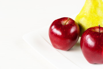 Red apples, green pear in white plate on white wooden background.