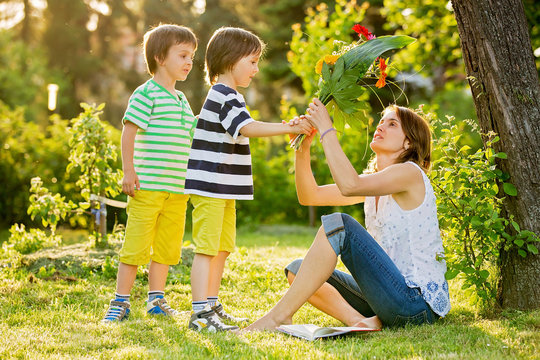 Young beautiful mother, sitting in a garden, little boys, her sons, bringing gerbera flowers and present