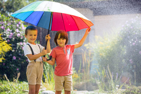 Two adorable children, boy brothers, playing with colorful umbrella under sprinkling water
