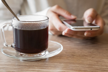Young woman drinking black coffee at the table in the cafe at lunch time. Hands holding a mobile phone and reading a text. Coffee break.