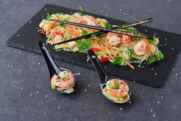 Noodles with prawns and vegetables on a black stone plate and in traditional soup spoons on a grey abstract background. Healthy food concept