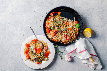 Whole-grain tomato pasta with basil pesto on cast iron pan with one white ceramic plate, lemon and garlic on textured rustic background