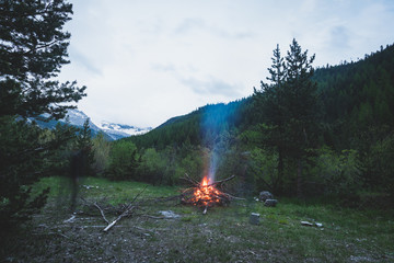 Burning camp fire into remote larch and pine tree woodland with high altitude landscape and dramatic sky at dusk. Summer adventures in the Italian French Alps.