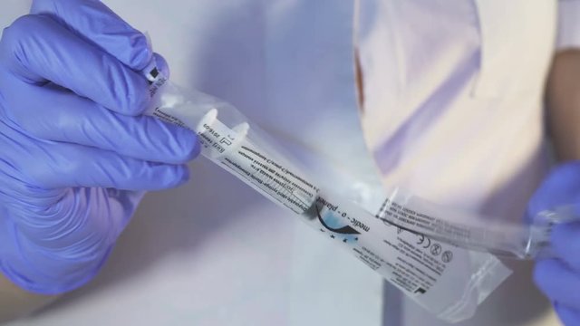 Nurse unpacks package with syringe while standing indoors. Clinic specialist in sterile blue gloves and white uniform cleverly opens transparent wrapping of miniature medical tool designed for