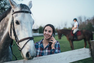 Beautiful young woman enjoying with her horse and talking on phone at ranch. 