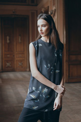 Portrait of young skinny model in top with birds print looking out the window in long corridor. Brunette pensive girl standing in classic interior. Fashion lookbook photo