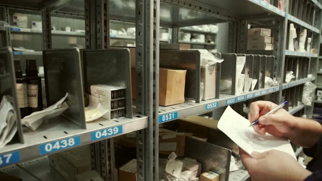 Warehouse worker makes a note in the documents and takes out a box. Inside are metal racks with numbered cells on shelves. Warehouse blue ballpoint pen makes a mark on the delivery note and pulls out