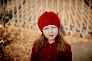 Baby 3 years with long hair. In a red beret and coat stands on the street in the sunshine, near the fence