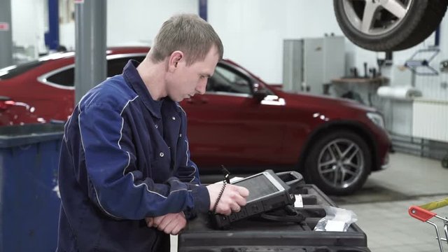 Mechanic standing at the bar and tap the screen to diagnose. He stands on the background of a red expensive car and looks carefully at the device. The professional is engaged in updating the software