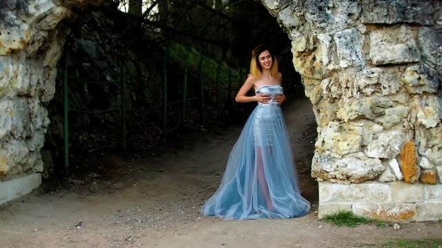Attractive girl with black brows and curly hair in silver and blue dress stands posing an smiling near stony wall of arс during photoshoot.