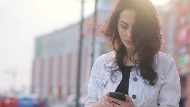 Attractive caucasian girl with serious face in white jeans coat and black t-shirt communicating with someone with smartphone and staying near huge shopping mall, shining sun, portrait in urbanistic