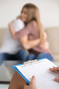 Close up of female psychotherapist holding clipboard, filling information about patients while couple embracing on couch in the background, doctor making notes in medical card in psychologists office