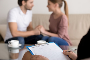 Successful therapy session with family couple in the background, reconciliation after argument, making peace, focus on hands of female psychologist holding clipboard with medical card, close up