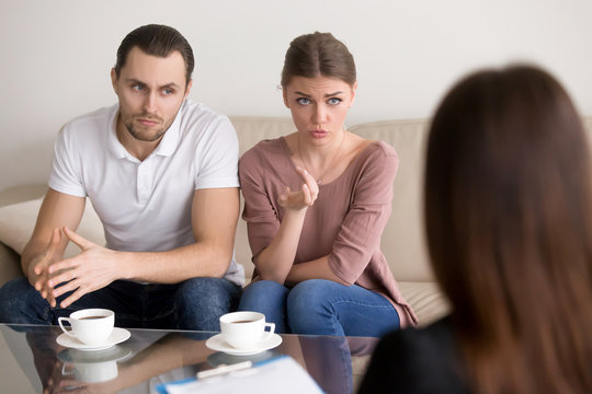 Portrait of beautiful serious couple sitting on couch and talking to psychologist. Young woman telling specialist about family problems, her husband sitting next to wife and listening attentively