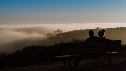 Fototapeta na wymiar Couple sitting on bench talking over a foggy sunset together, relationship