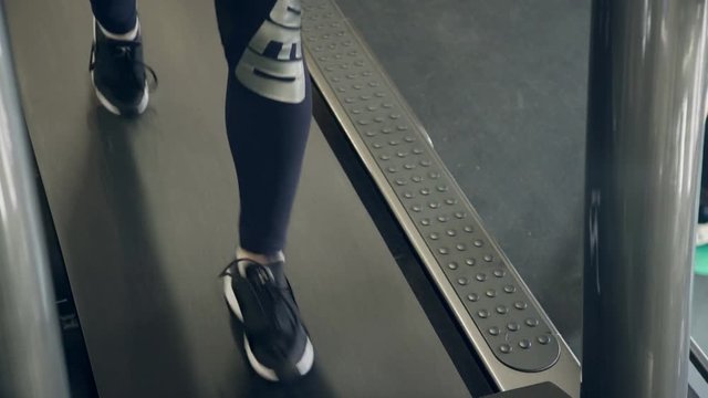 Young athlete walks along treadmill in gym before training. Lady dressed in compression pants and black sneakers going fast along moving racetrack. Early saturday morning she came to sports club for