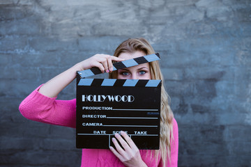 Happy young blond woman holding cinema clapper board and smiling over gray concrete or brick wall...