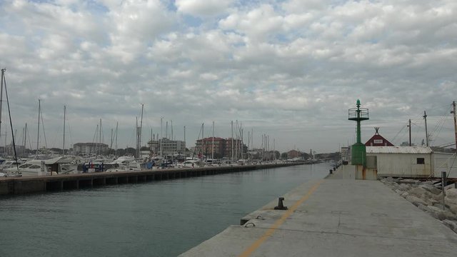 bollards and steel lighthouse near fishing hut on pier along sea channel while boats and ships are moored in the marina