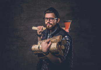 Bearded male in eyeglasses dressed in fleece shirt holds firewoods and axe on his shoulder.