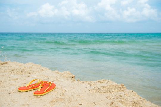 Sandals on the beach, summer holiday vacation concept. Orange sandals with turquoise sea in the background with cloudy sky.